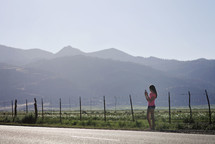 a teen girl standing on the side of a rural road taking a picture with her cellphone 