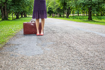 legs of a woman on a rural road next to a suitcase 