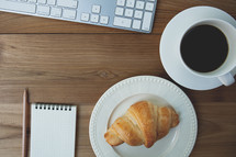 croissant on a plate, notepad and pencil, computer keyboard, and coffee mug on a table 