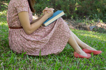 a woman in a pink dress sitting in the grass praying over a Bible 