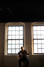 man playing a guitar in front of a window 
