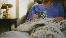 woman under blankets with a coffee mug and book 