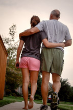 A father and daughter walking down a sidewalk with arms around each other.