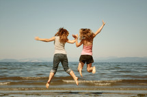 Two girls holding hands and jumping into the air on the beach.
