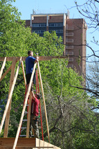 Construction workers putting up roof joists with trees and a tall building in the background.