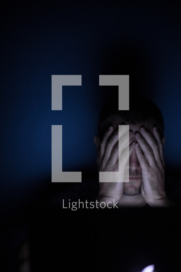 a man covering his eyes looking at a computer screen at night 