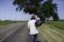man standing in the middle of a road with his back to the camera carrying a bag 