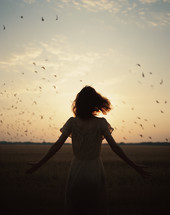 A woman standing in a field with outstretched arms during sunset with birds in the background