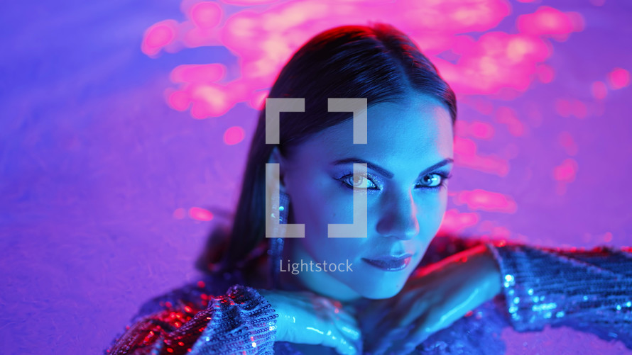 Beautiful woman posing in pool water under neon color light. Party, attractive chic in shining dress enjoying night time. Rich lifestyle, luxury life, influential people