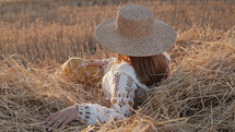 Pretty woman in straw hat and embroidered blouse smiling lying on hay in countryside at sunset. Rural nature, haystack, vacation, relax and harvest concept.