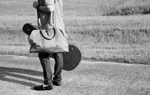 a man standing on a road carrying a duffle bag and guitar case 