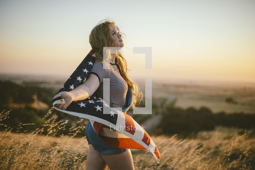 a teen girl carrying an American flag in a field at sunset 