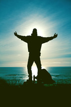 Silhouette of a man standing with outreached arms at the ocean with an aura of light around him.