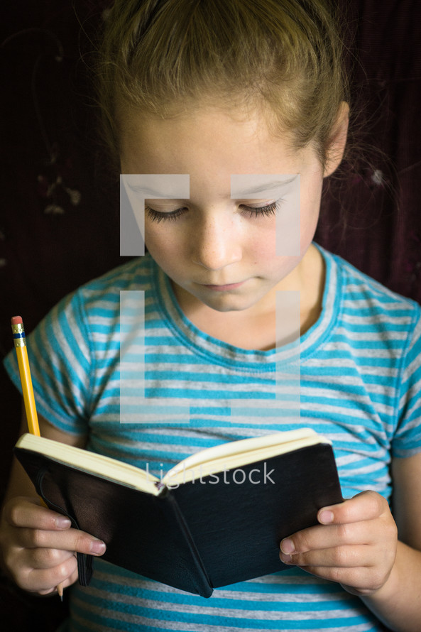 A girl reading a notebook and holding a pencil.