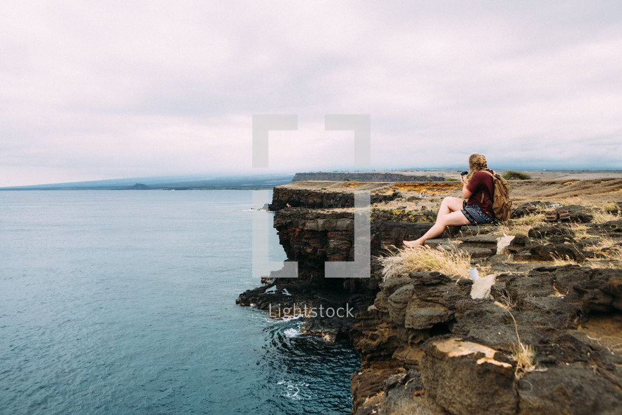a woman sitting on a cliff at a shore looking out over the ocean 