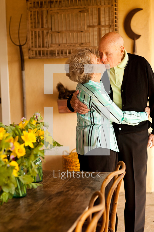 Grandparents embracing and kissing in a sunroom.
