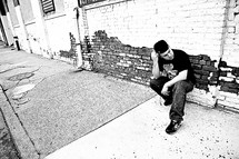 man crouched on a sidewalk in front of a brick wall