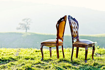 Two antique chairs in a grassy field  facing away from each other on a hillside