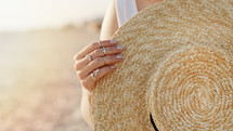 Woman hand with boho gypsy rings holding straw trendy hat. Girl standing on sandy beach at summer near sea and rolling her boater. High quality photo