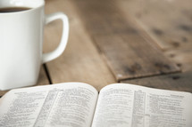 pages of an open Bible and coffee mug on a wood table 