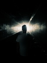 A man walking through a dark tunnel toward a bright light.