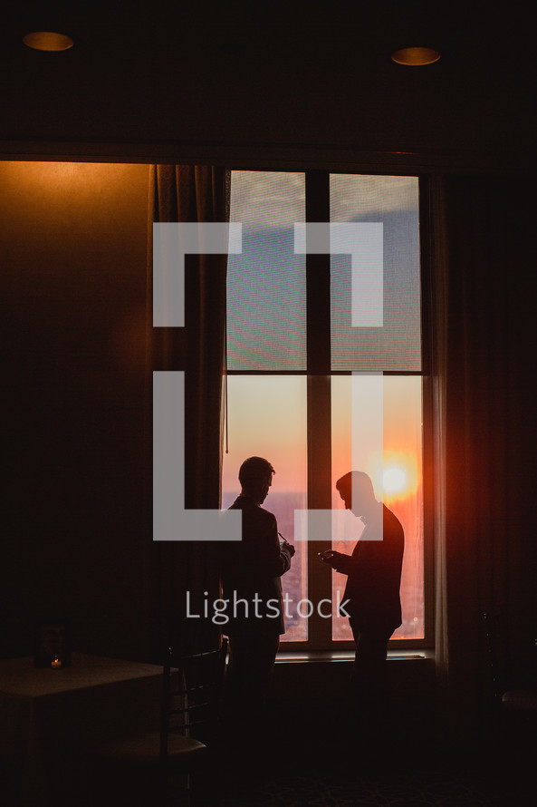 men standing by a window at sunset looking at their cellphones 