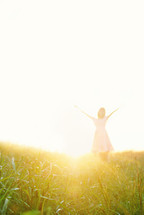 Hazy sunlight on a little girl standing in a field of grass with her arms raised toward the sky.