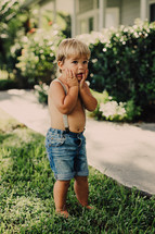 a bare chested toddler boy in suspenders standing in the grass 
