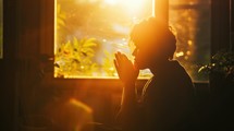 Silhouette of young man praying in front of window at home.