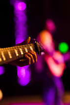 Hand holding guitar with other player in background on lighted stage fret board