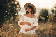 Happy Mother Carrying Child, Expecting For Baby. Amazing Motherhood. Pregnant Woman in beautiful dress and straw hat sitting on grass at nature rural meadow. High quality 