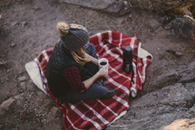 a woman sitting on a plaid blanket outdoors drinking coffee