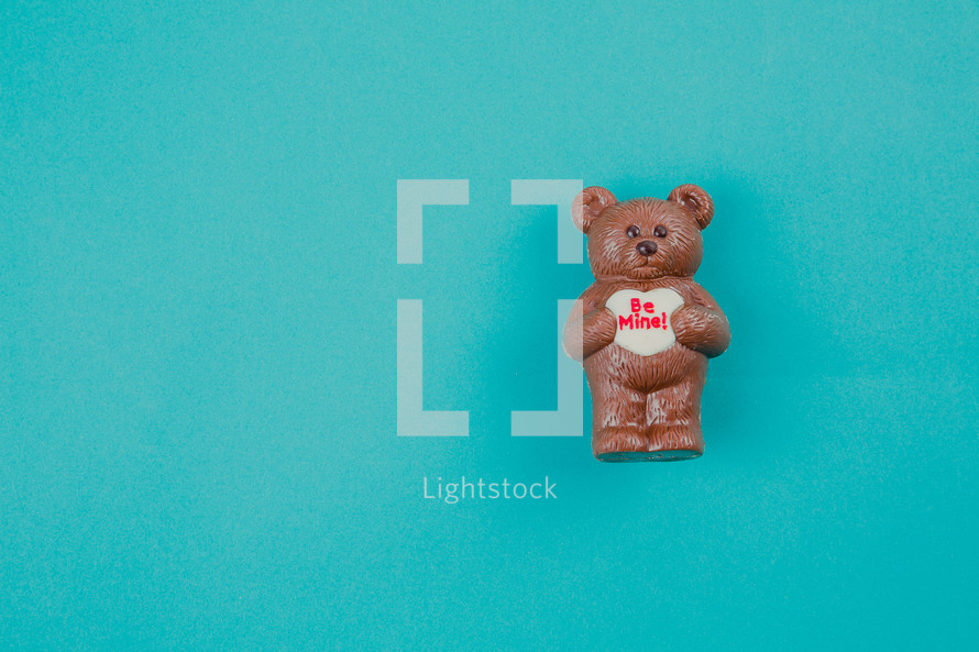 A chocolate bear holding a heart that says "be mine" on an aqua background.