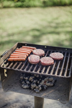 Hot dogs and hamburgers cooking over hot coals on an outside grill.