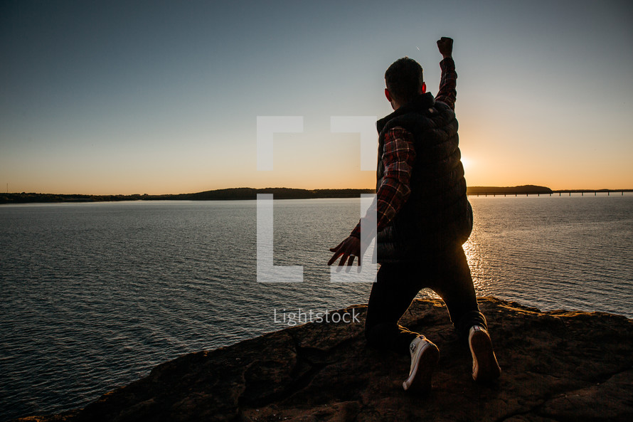 a person kneeling with a raised fist at the edge of a cliff at sunset 