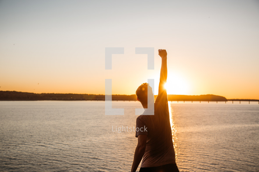 a person standing with a raised fist at the edge of a cliff at sunset 