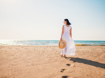 Girl wearing white maxi long dress walking barefoot on the sea shore with hat in hands. Bohemian clothing style.
