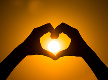 Hand heart frame shape silhouette made against the sun and sky of a sunrise or sunset on a deserted empty beach
