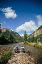 men sitting in folding chairs along a rivers edge near a campsite 