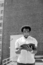 An African American man standing reading a Bible in a city 