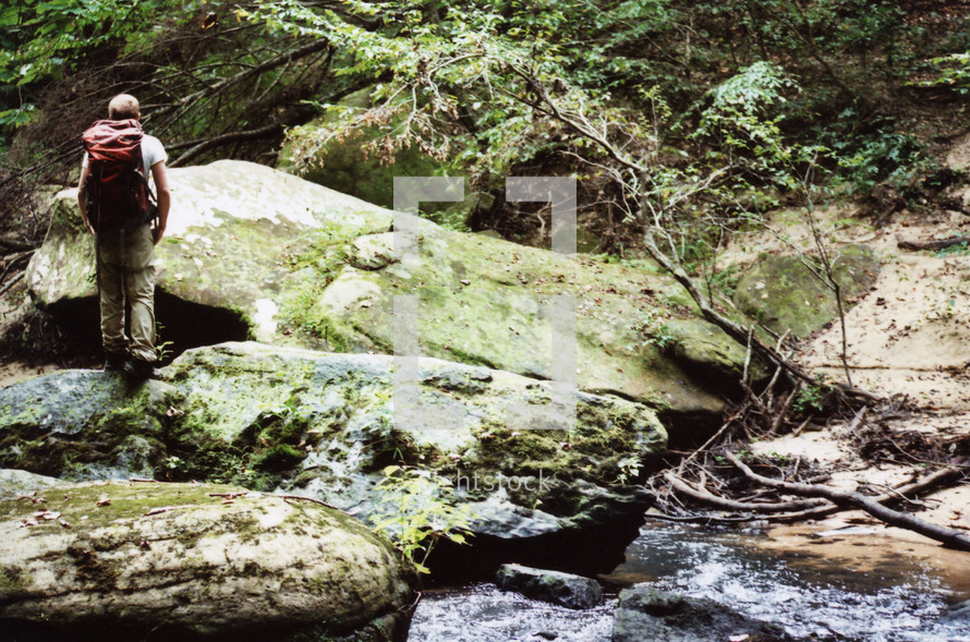 man standing on moss covered rocks looking at a stream 