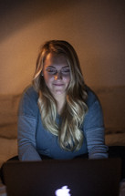 young woman sitting on her bed looking at a computer screen in a dark dorm room 