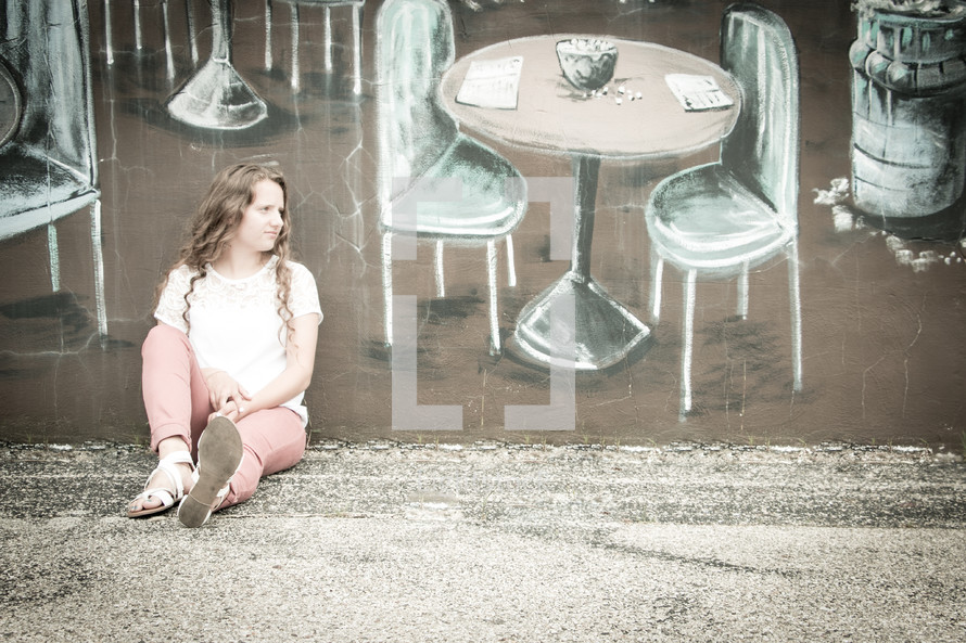 woman sitting on a sidewalk in front of a painting of table and chairs on a wall 