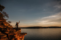a person kneeling with a raised fist at the edge of a cliff at sunset 
