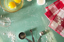 whisk, flour, measuring spoons, cracked eggs, bowl, and towel on a kitchen countertop 
