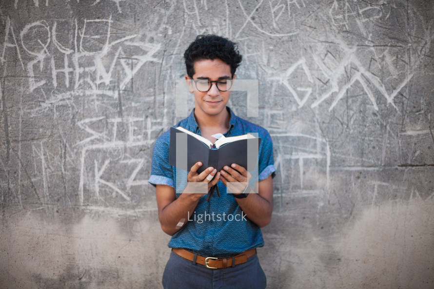 man reading a Bible in front of a gray wall with graffiti 