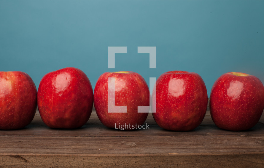A row of bright red apples on a wooden surface against a blue background.