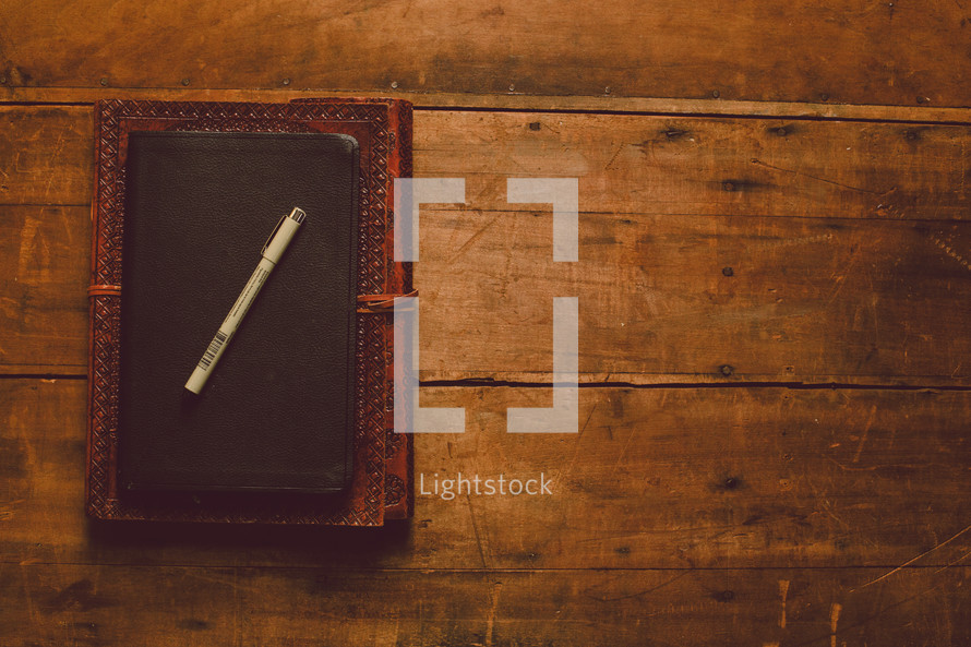 A leather journal, Bible, and a pen stacked on a wooden table.