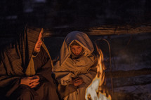 A man and child dressed in Biblical clothing sit near a fire.