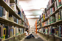 student sitting on a floor of a library reading a book.